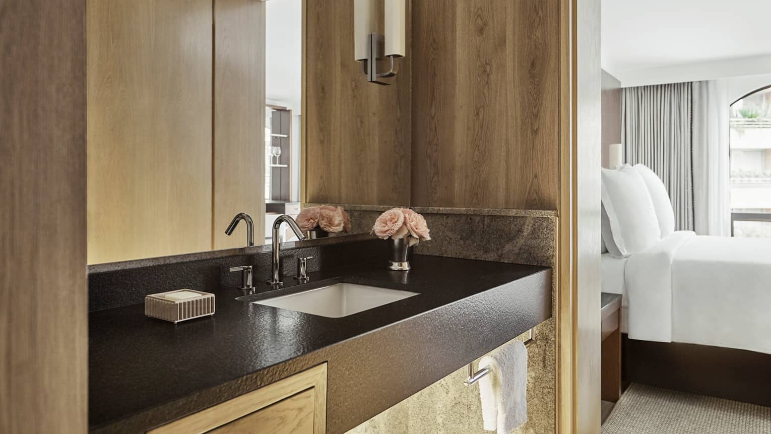 Close-up of bathroom vanity with black counter, sink, wood walls, bedroom visible to side