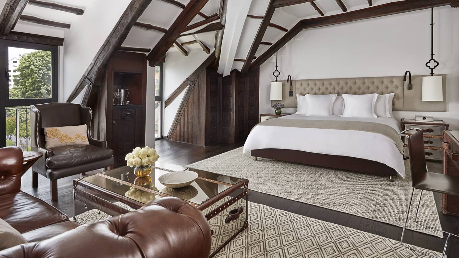 Sloped beam loft ceiling over hotel bed with padded headboard, brown leather armchair, table with flowers