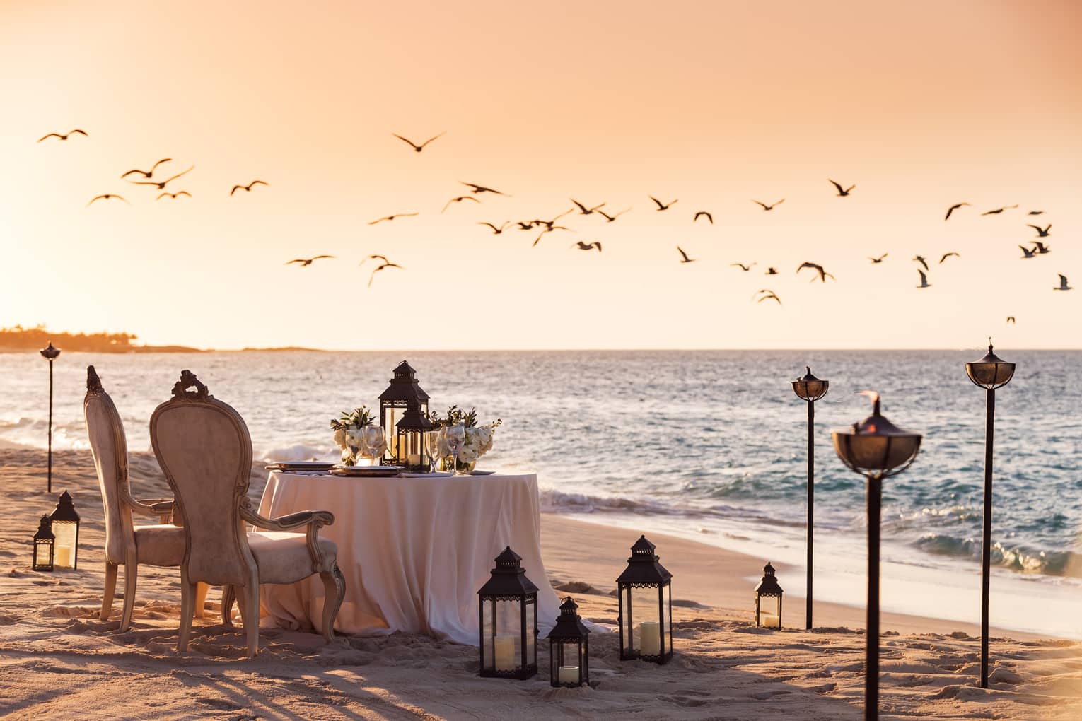 Romantic beachside dining setup with vintage chairs, a table decorated with flowers and lanterns and birds flying overhead