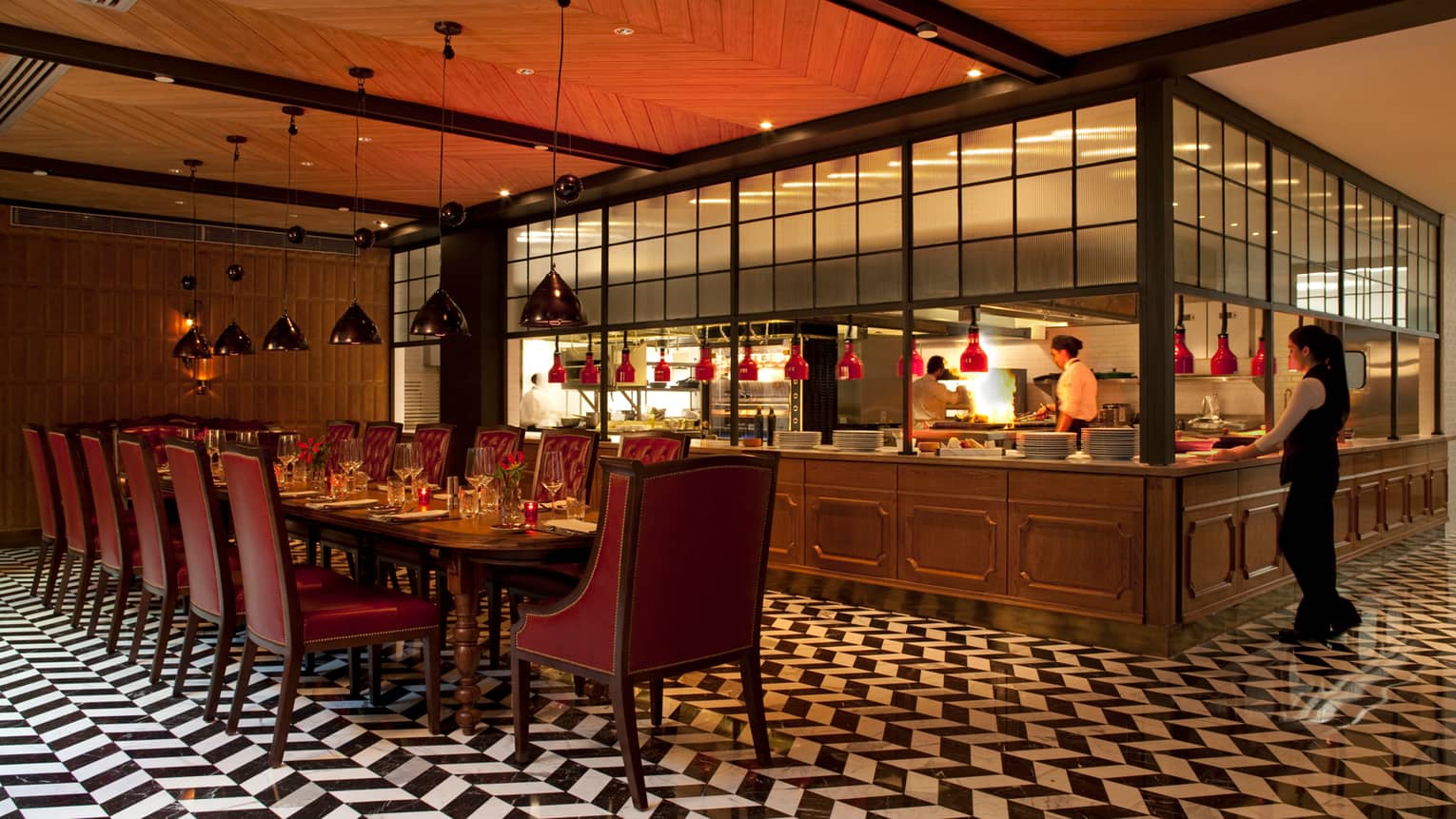 Long 14-person dining table with red leather chairs in front of wood kitchen line, server, black-and-white tile floors