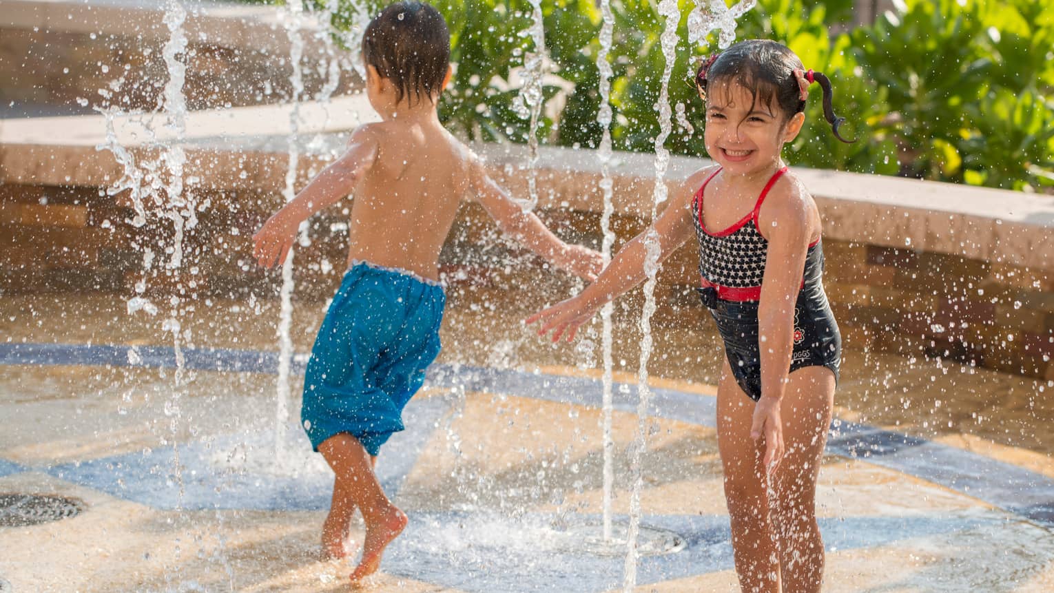 Young boy and girl in swimsuits run through water splash pad 