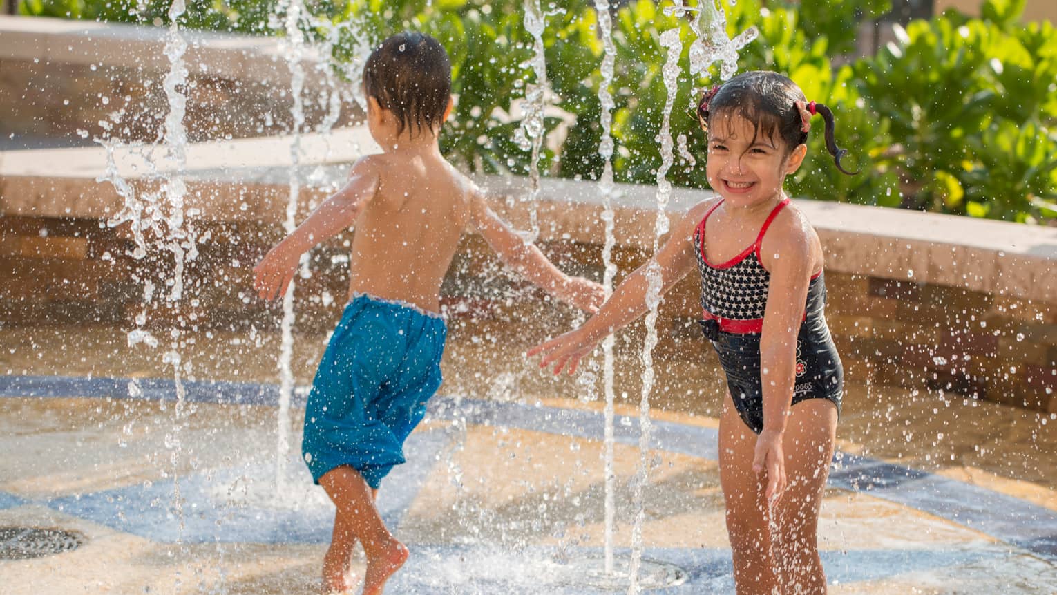 Young boy and girl in swimsuits run through water splash pad 