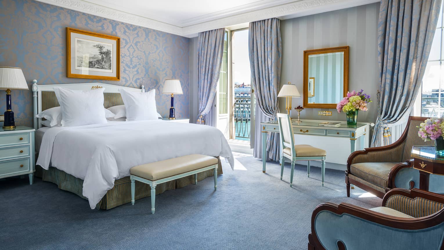 Luxury hotel room with a king bed, nightstands with lamps, seating area, desk and a balcony with light blue and gold decor