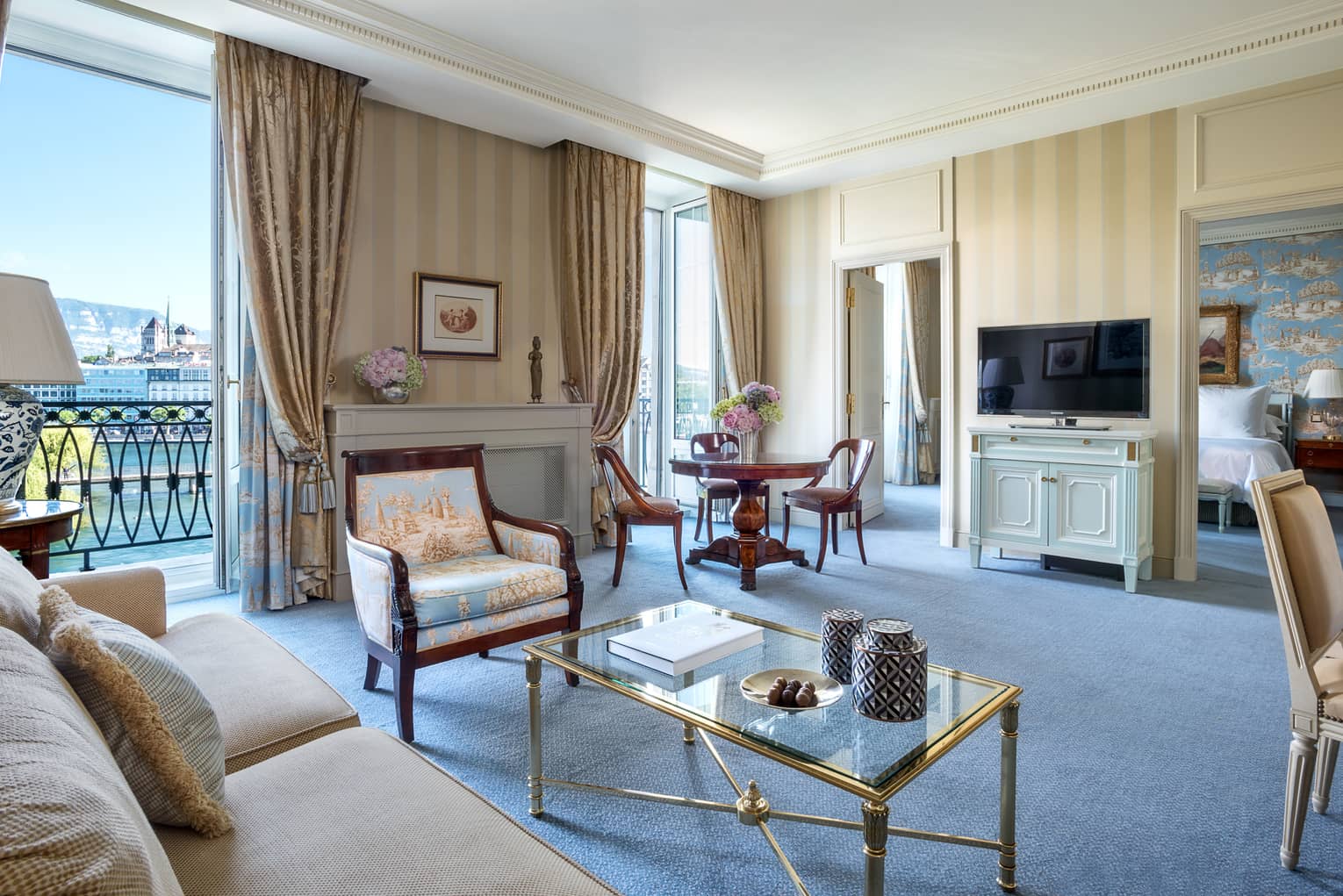 Spacious hotel suite with a beige sofa, glass coffee table, antique-style armchair, a round dining table, striped wallpaper, TV and a balcony with a lake view