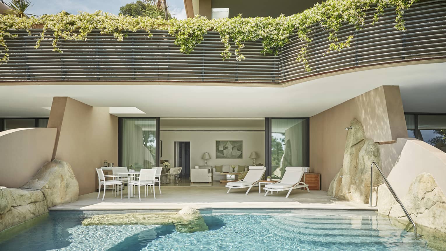 Hotel suite patio with white chairs beside private pool flanked by boulders and view into living room