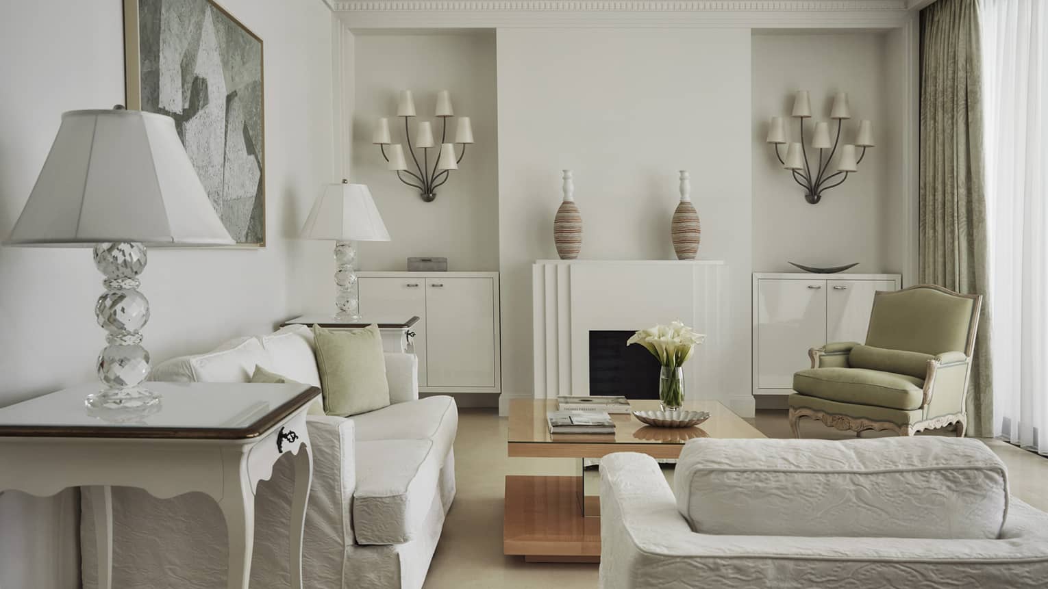 Hotel suite living room with white sofa and club chair, coffee table and fireplace flanked by wall-mounted light fixtures