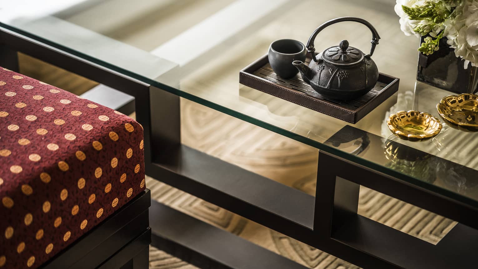 Close-up of glass table with black teapot set on tray, gold decorative dishes and red cushioned seat in luxury hotel setting.