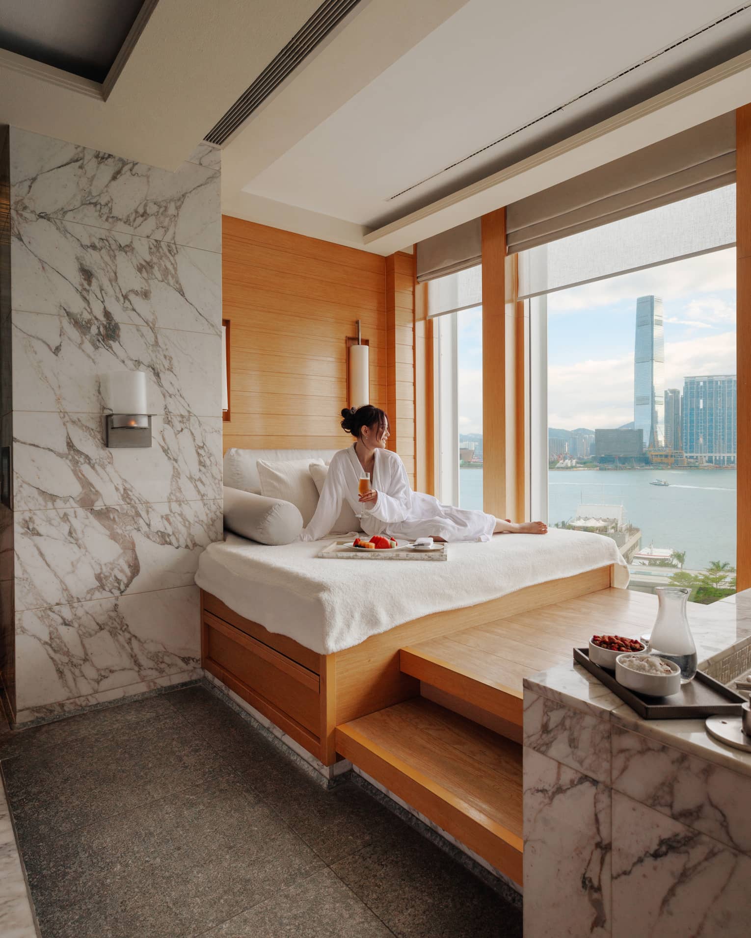 Woman in spa bathrobe sits on windowside chaise looking out to Victoria Harbour at Four Seasons Hotel Hong Kong