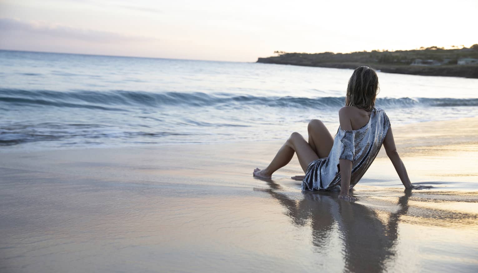 View from behind of a guest sprawled out on wet sand, gazing out at pale pink and buff beginnings of sunset over calm ocean.