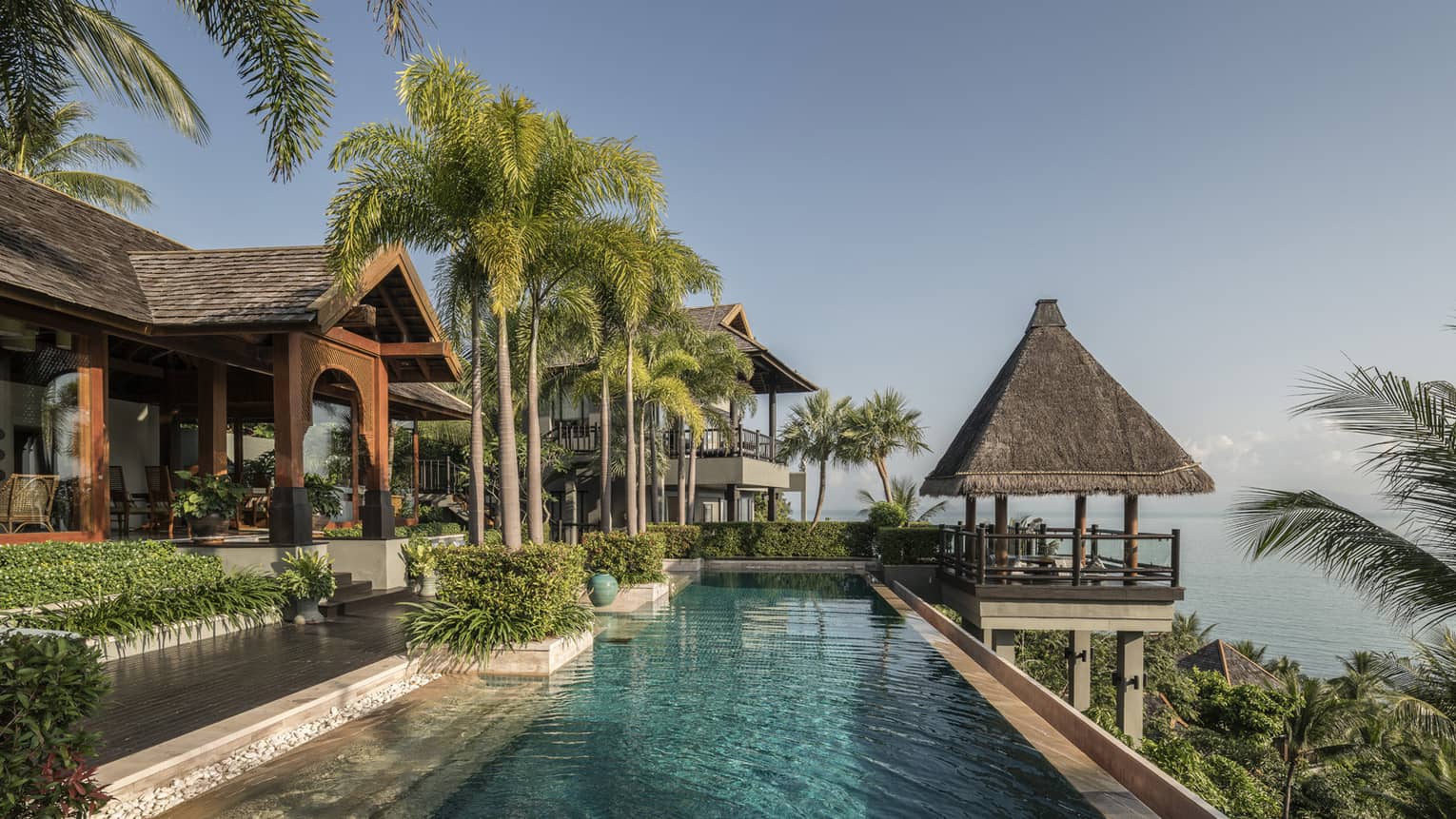 Long outdoor swimming pool under tall palm trees, thatched roof gazebo