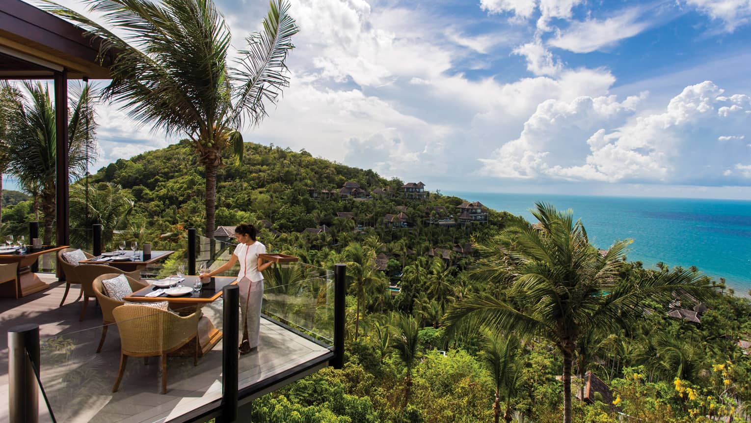 Server sets corner patio table behind glass balcony, high on tropical mountain