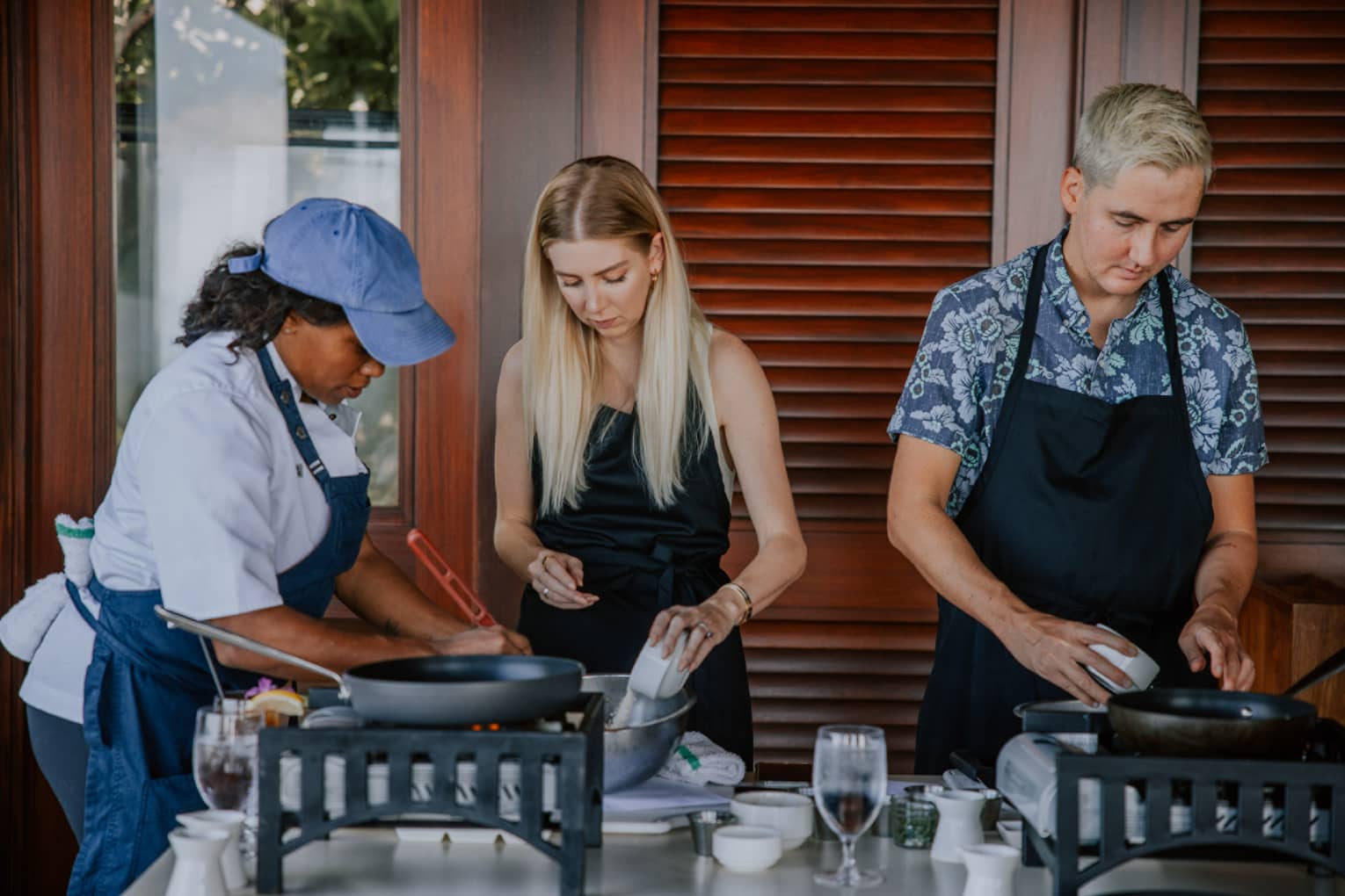 A cooking class is underway as three people sporting aprons stand over a table strewn with wine glasses and cookware.