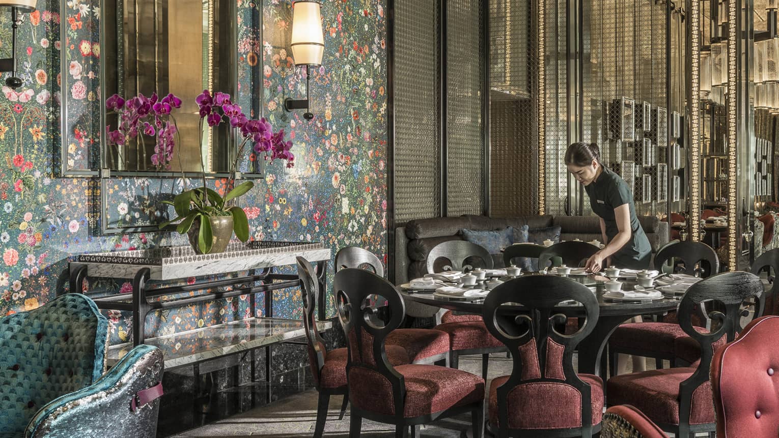 Hotel staff sets table with elegant red velvet chairs under tall mirror, floral wallpaper 