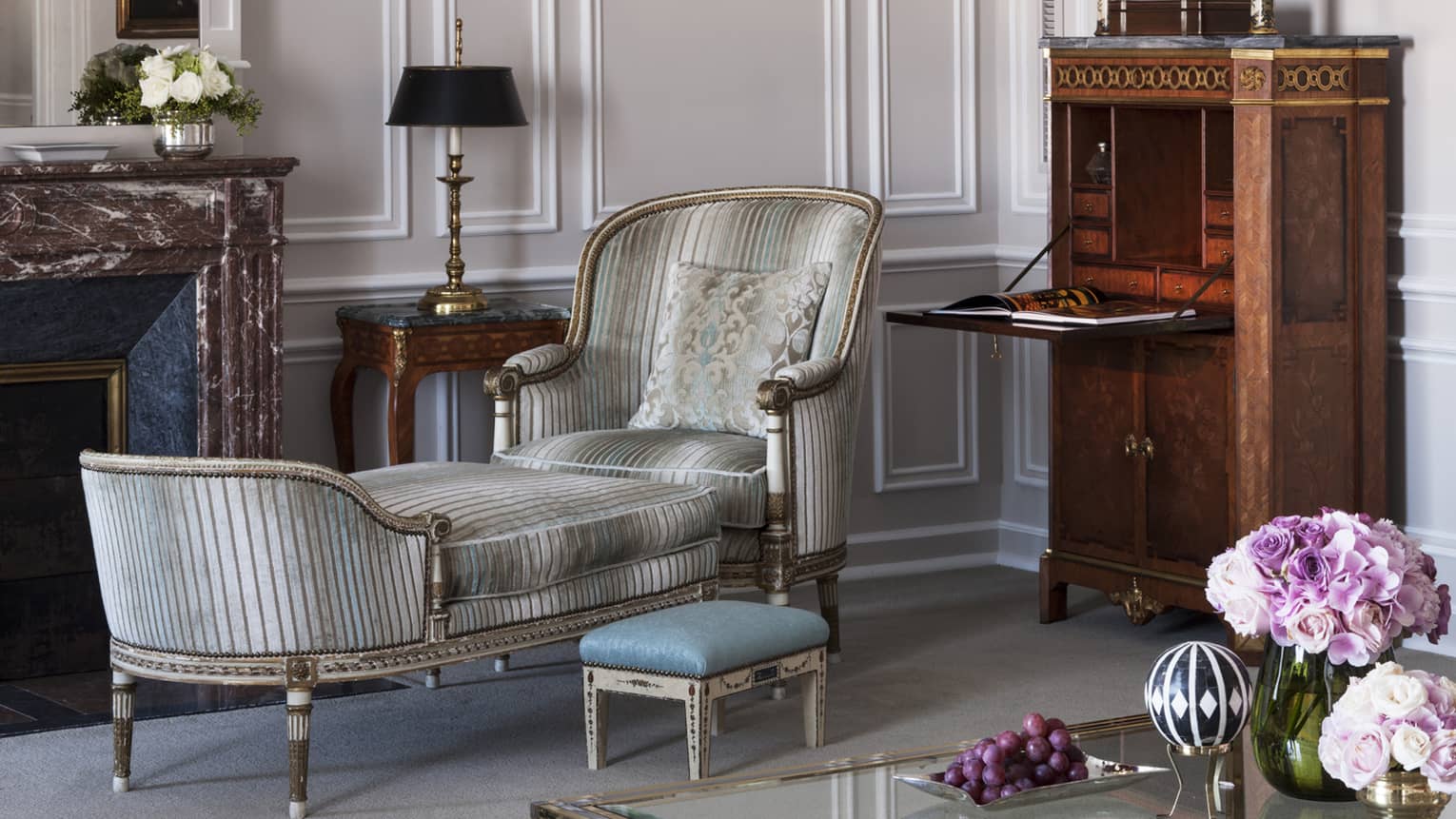 An elegant living room with a vintage striped lounge chair, an ornate marble fireplace and a wooden secretary writing desk. The decor includes floral arrangements and classic artwork.