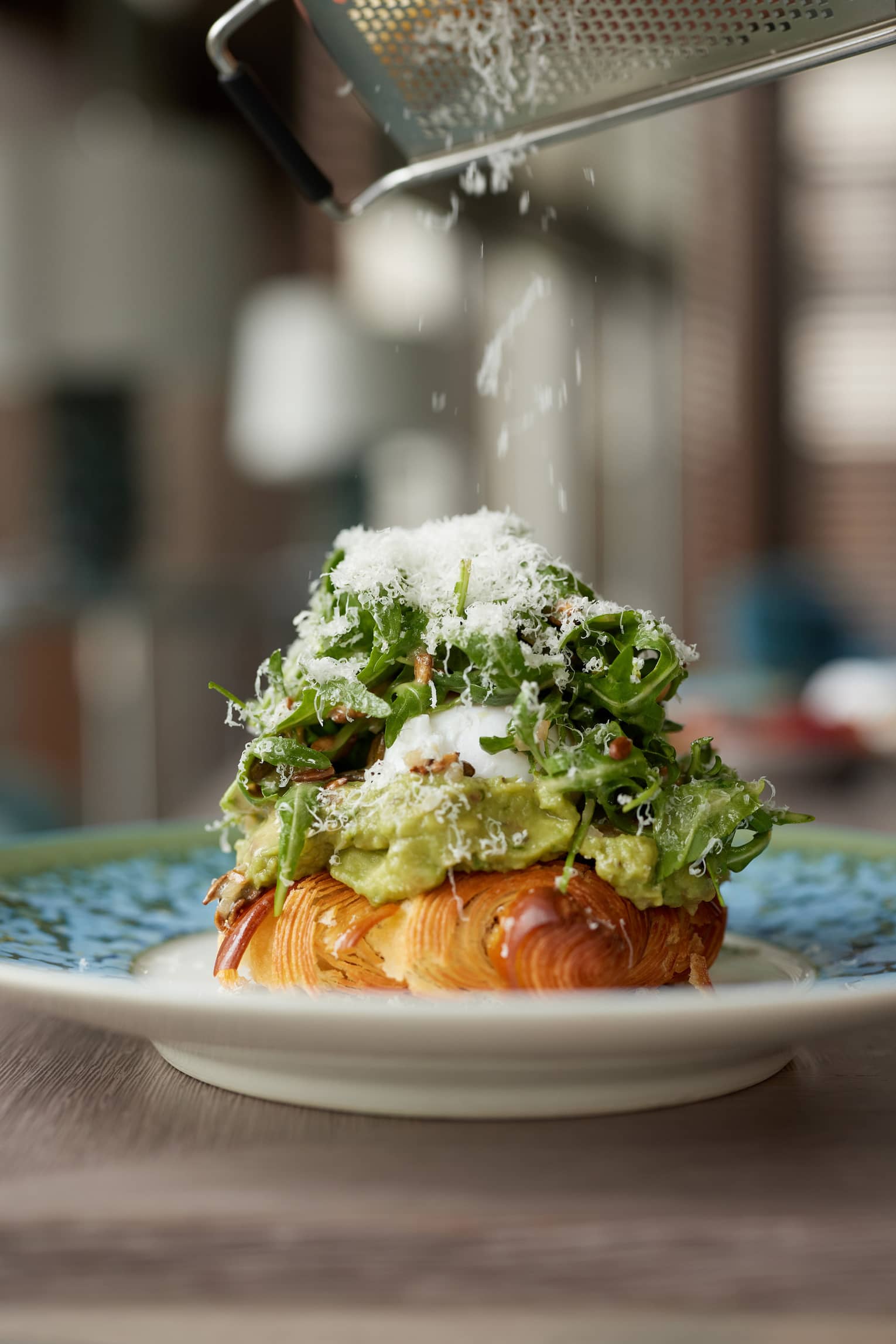 A close-up of a plate holding a croissant covered with avocado, egg and arugula salad topped with grated parmesan. 