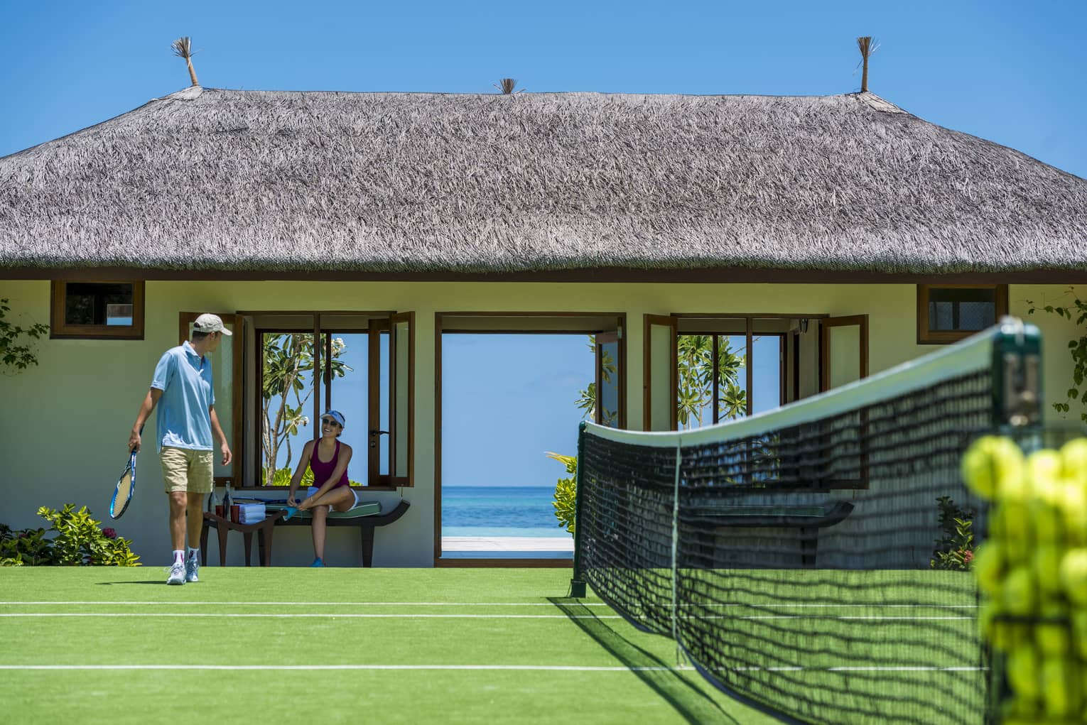 Two guests prepare to play tennis on a court bordered by a grass-roofed building with doors opening onto a view of the ocean.