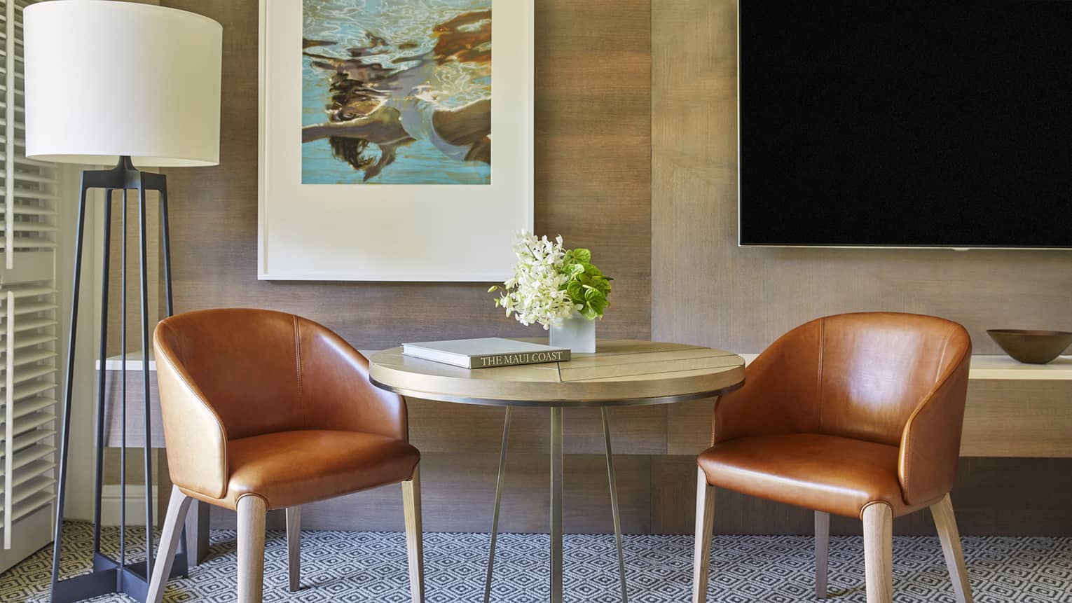 Seating area with two brown leather chairs, round wooden table with flowers, floor lamp, wall art and mounted TV