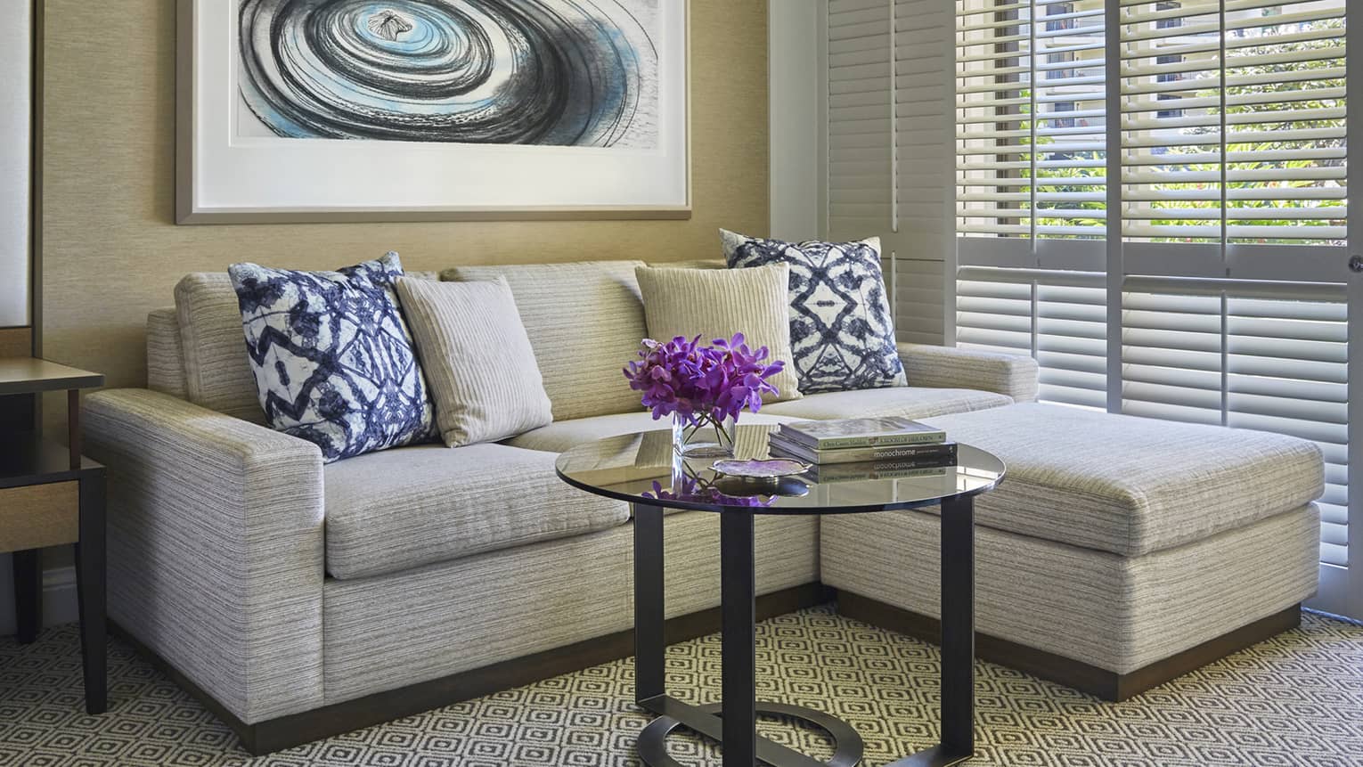 Living area with beige sectional sofa, pillows, round glass coffee table with purple flowers and abstract wall art