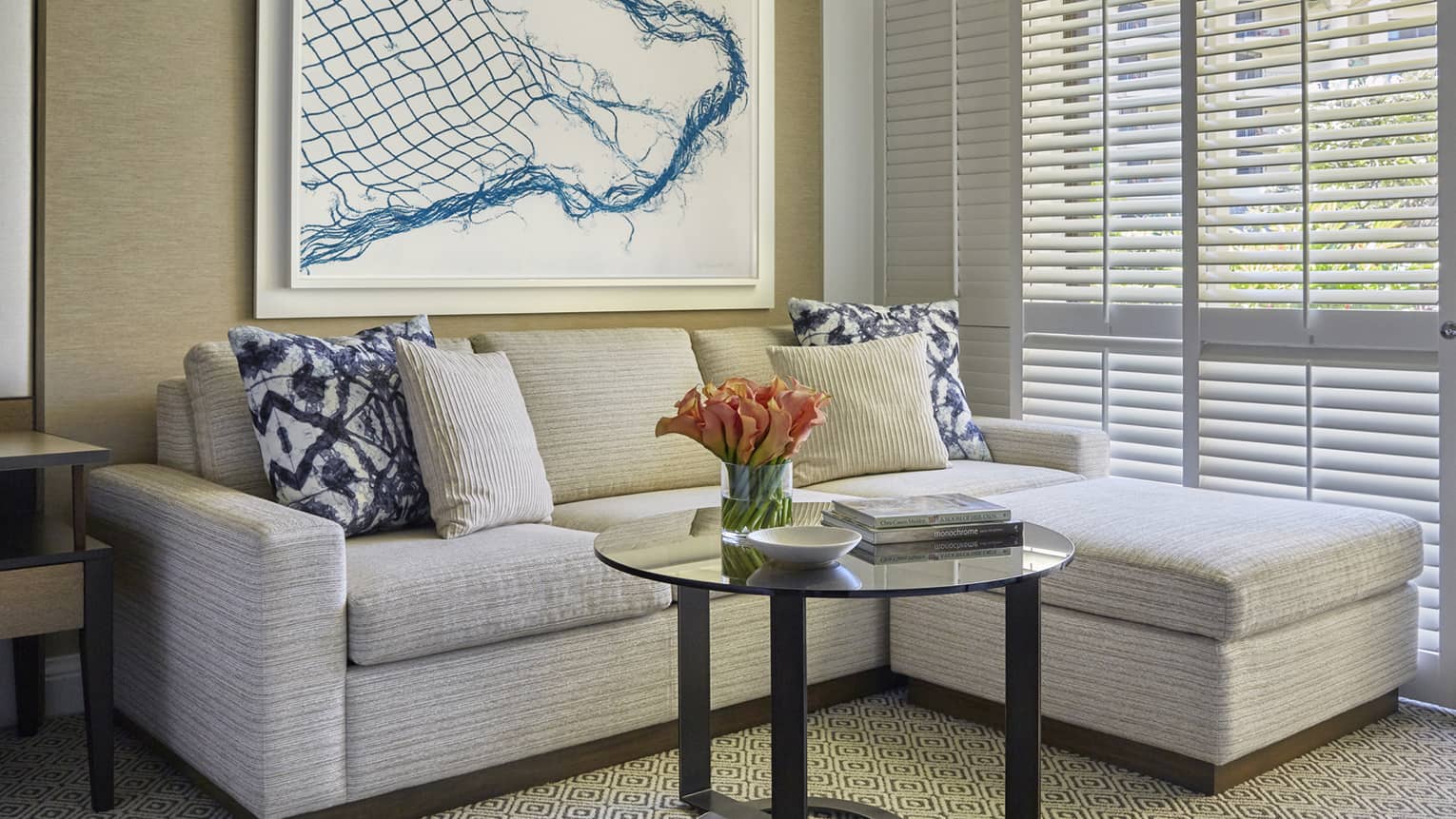 Cozy seating area with beige sectional sofa, blue and striped pillows, glass coffee table with orange flowers and abstract wall art