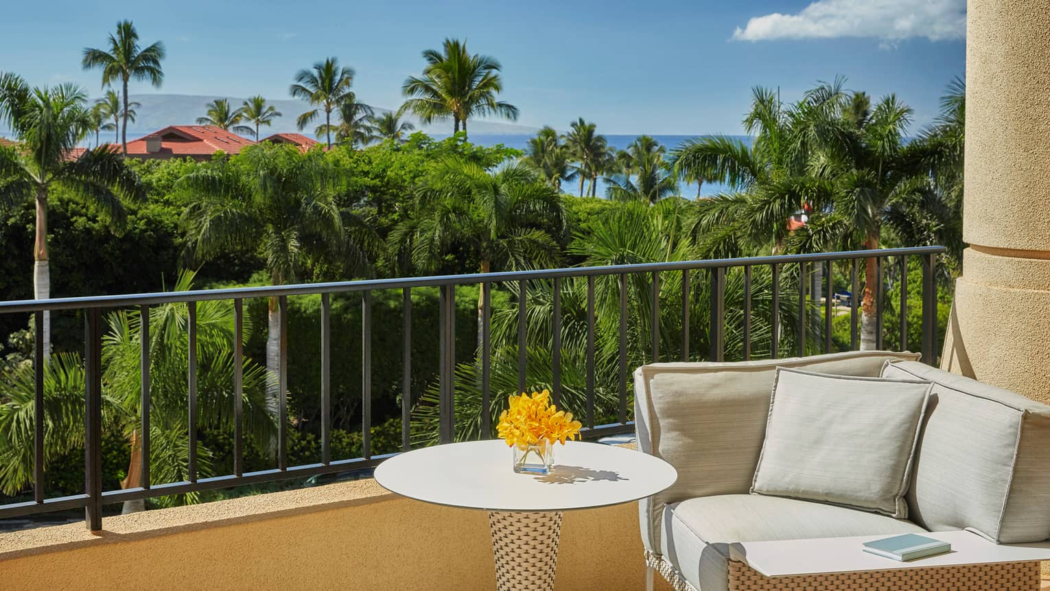 Resort balcony with white table holding yellow flowers, cushioned seating and view of lush tropical greenery and ocean