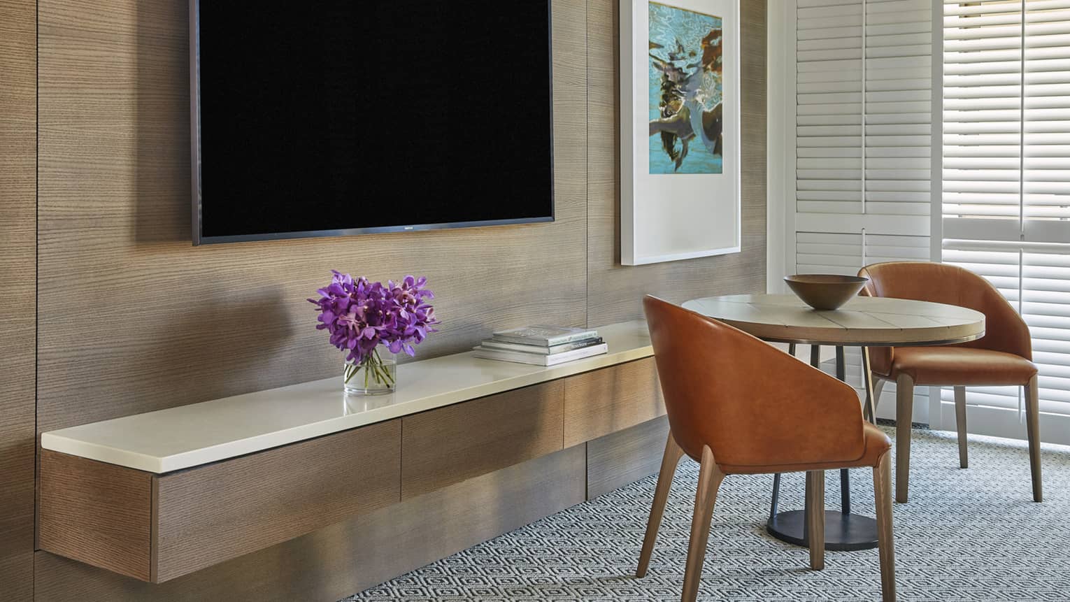 Resort room area with wall-mounted TV, floating console with purple flowers, round table and two brown chairs