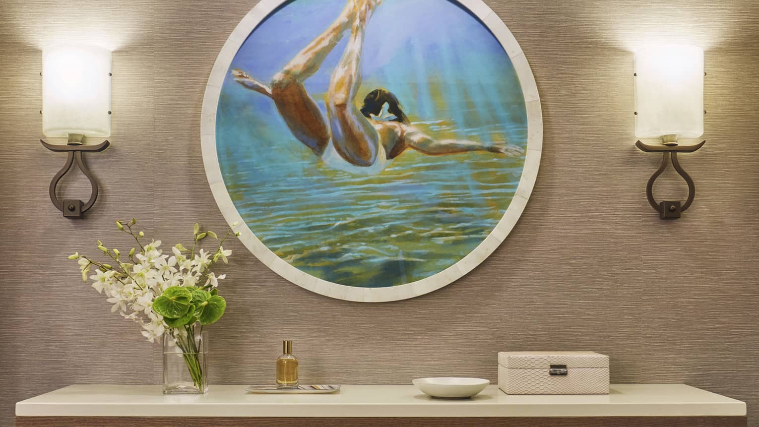 Wall art of a swimmer above a table with flower arrangement, small perfume bottle and decorative items, flanked by two wall sconces