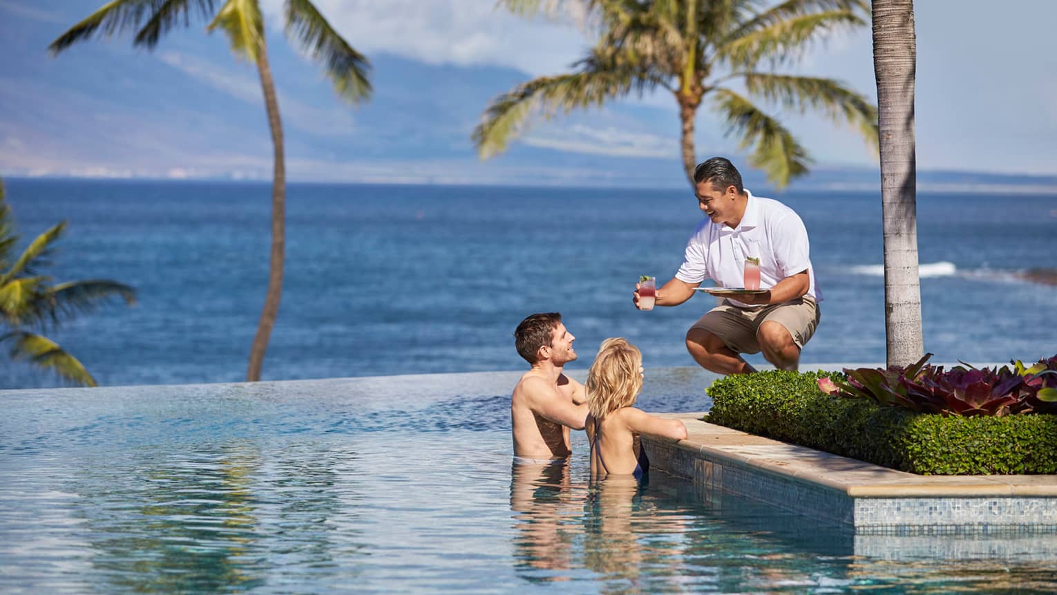 Server in white uniform with tray passes a cocktail to man and woman at edge of infinity swimming pool