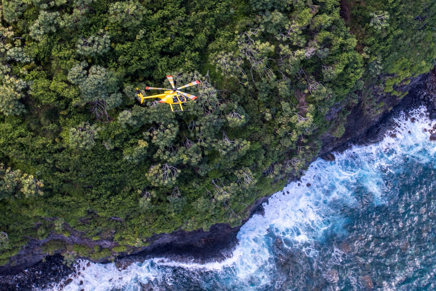 A yellow helicopter flies over a steep forested mountain. Powerful waves crash against the shoreline in the water below.