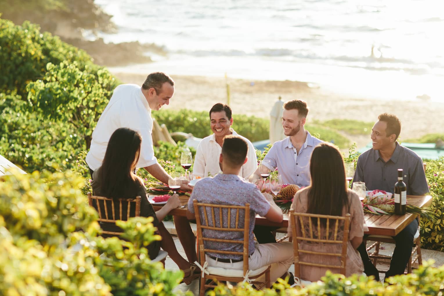 Six people smiling, sit around outdoor wooden table by ocean as wait staff serves them