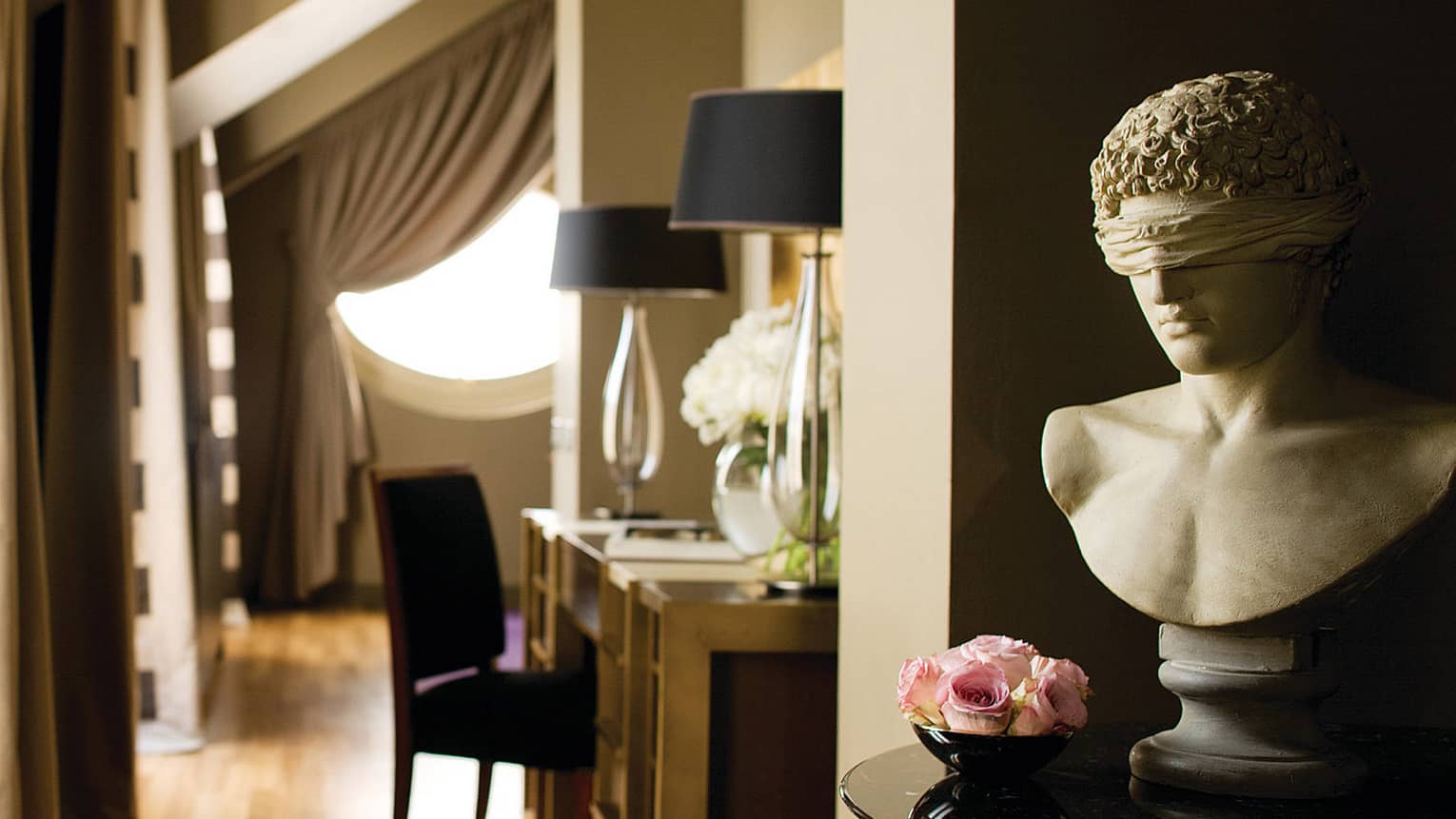 Hotel suite desk with purple chair topped with glass lamps, windows behind flanked by striped curtains, blindfolded bust in forefront