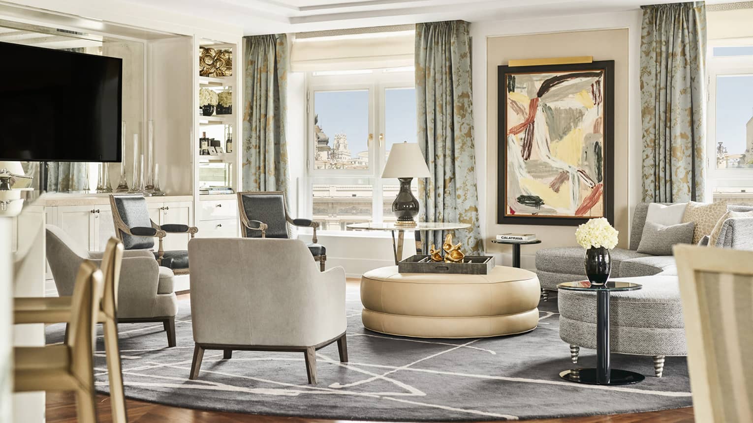 Hotel suite with abstract artwork, TV, grey armchairs and sofa, round beige ottoman, modern decor, glass tables and floor-to-ceiling windows with city views