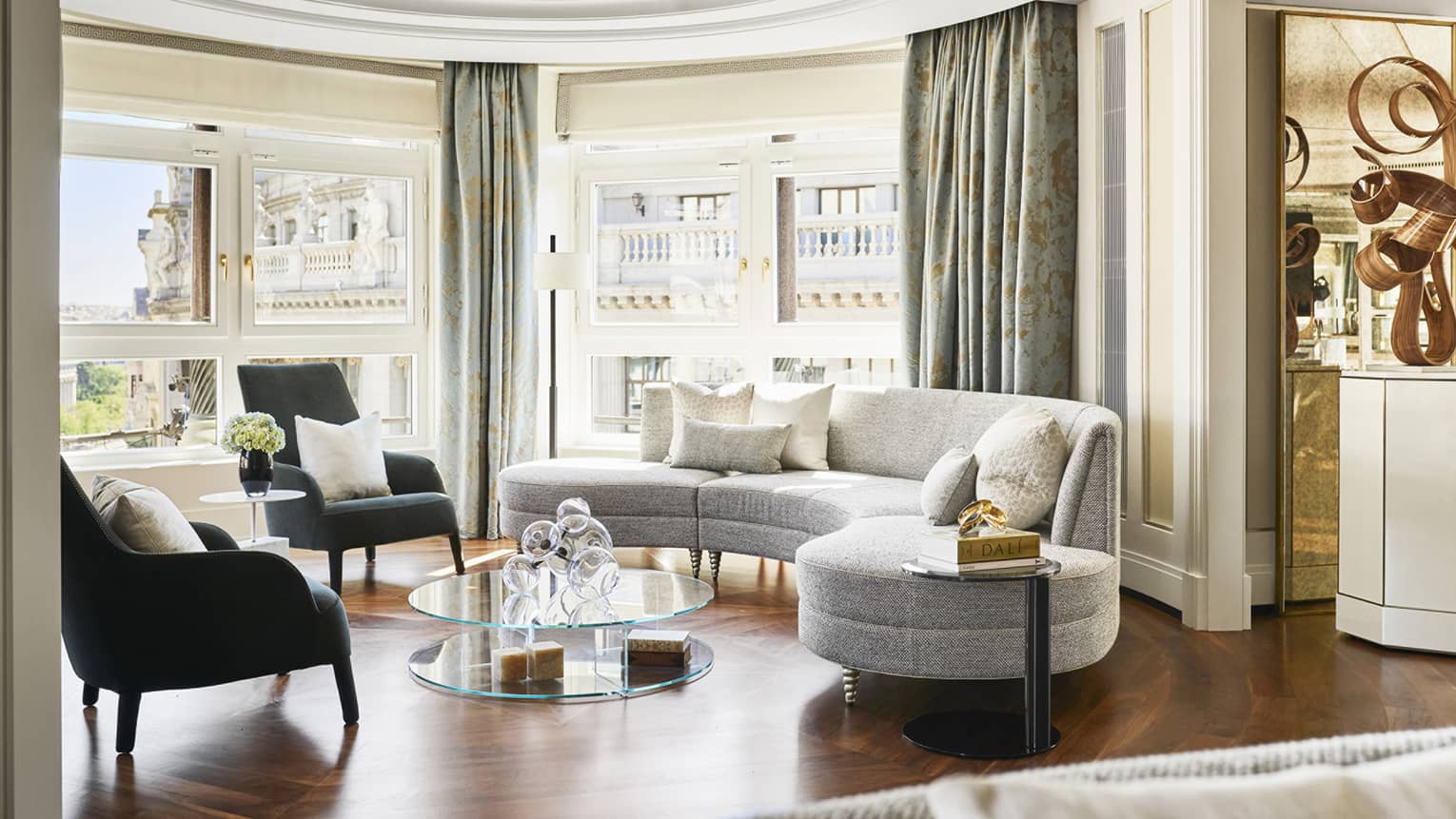 Luxury hotel suite with curved grey sofa, armchairs, round glass coffee table, floor-to-ceiling windows and city views framed by patterned drapes