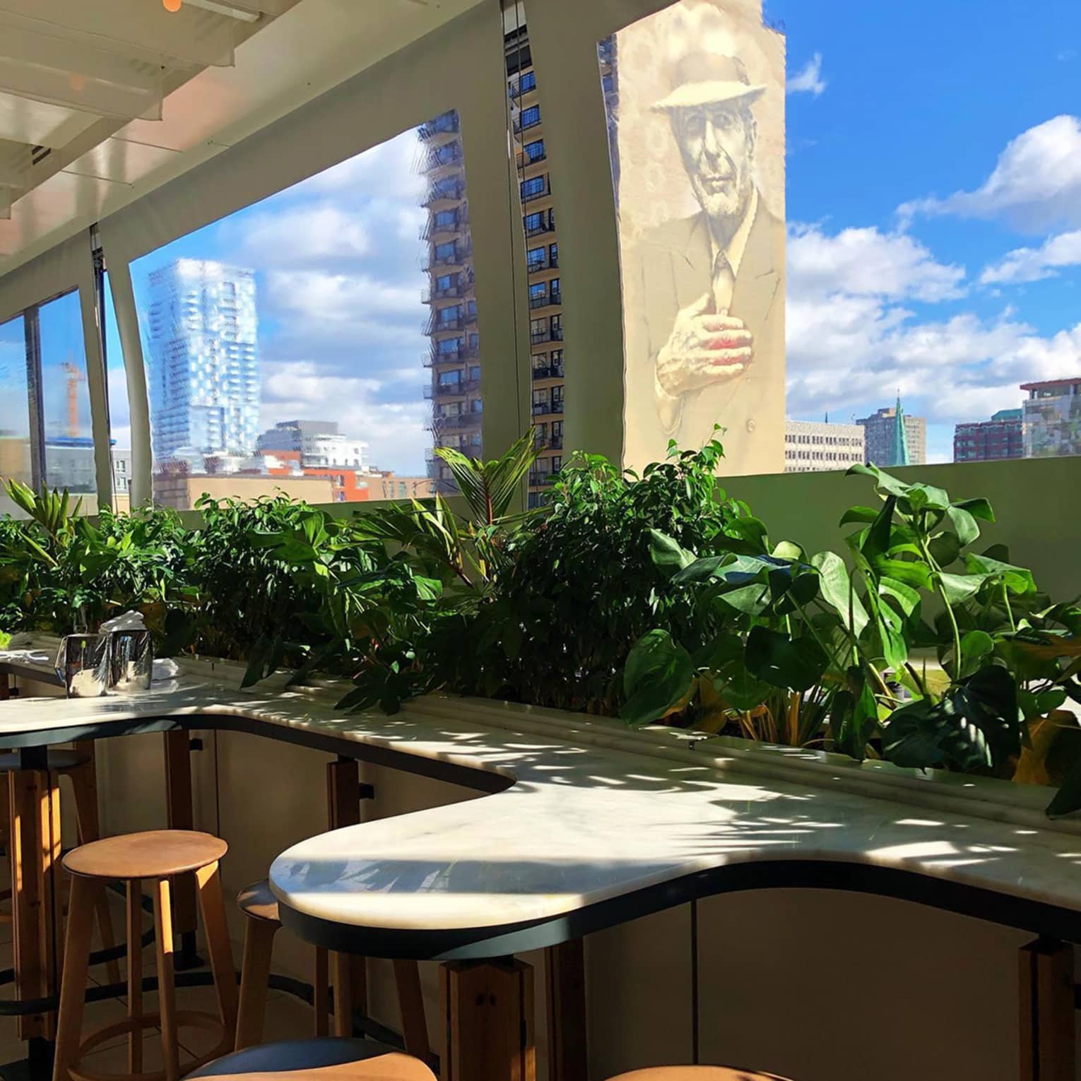 A bright café boasts curved marble countertops and wooden stools, featuring lush greenery. Large windows offer views of city buildings, including a mural of a man in a hat.