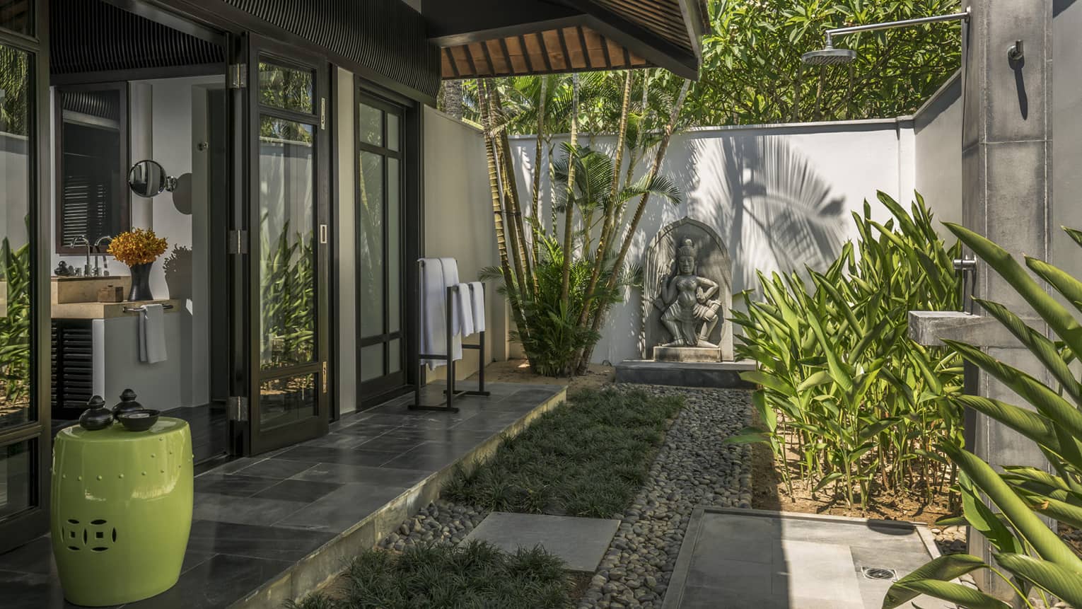 Outdoor shower area with a tropical garden, stone statue, and modern fixtures connected to a villa's bathroom through glass doors.