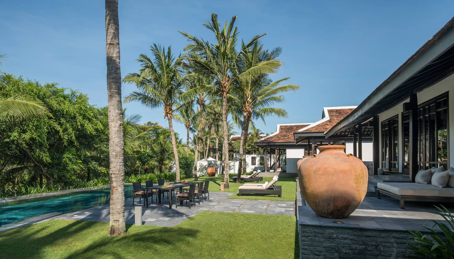 Luxury villa garden with a swimming pool, outdoor dining area, sun loungers and large clay pots, surrounded by palm trees and lush greenery under a clear sky.