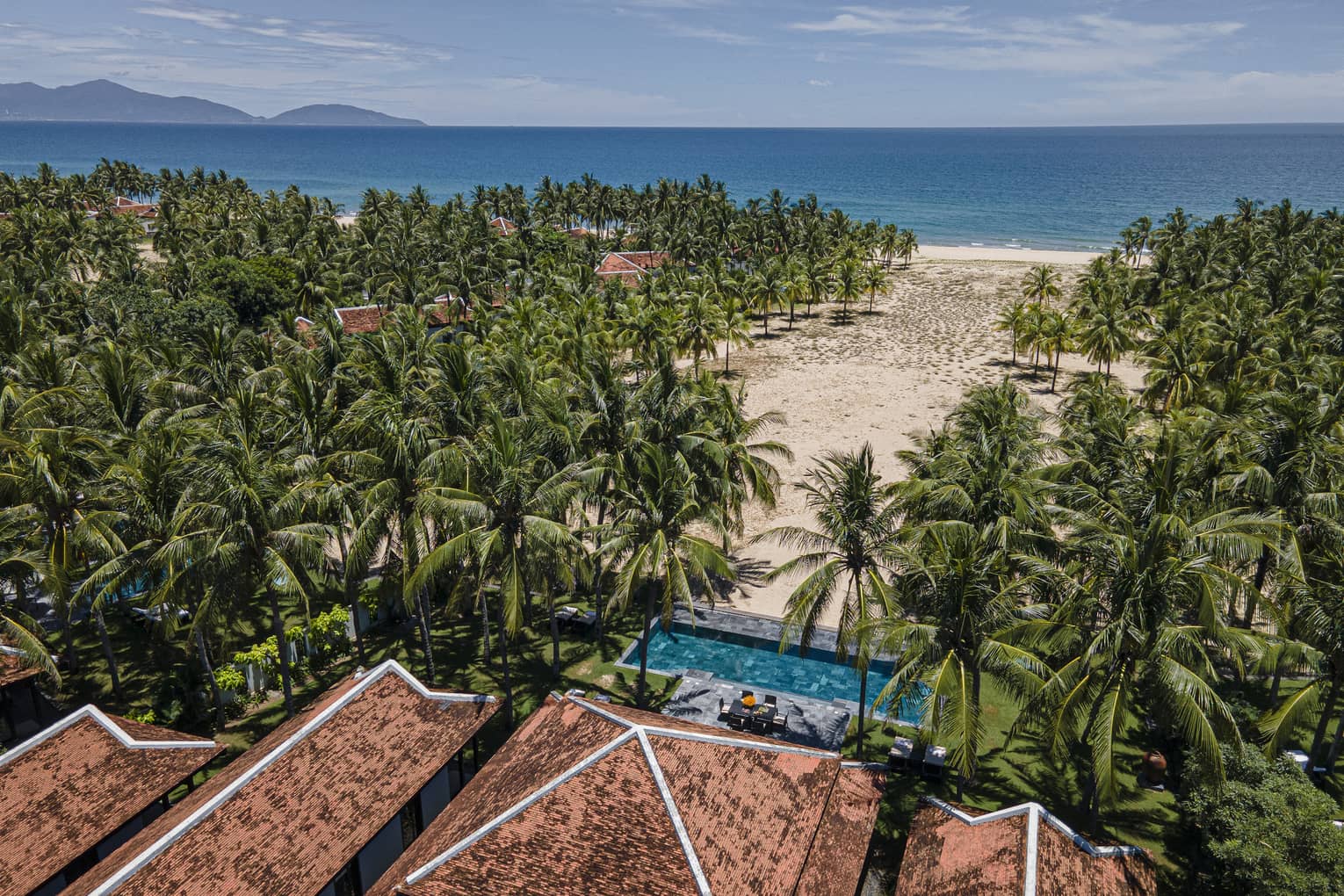 Aerial view of a beachfront Resort surrounded by lush palm trees, with a sandy beach, turquoise ocean and distant islands under a clear sky.