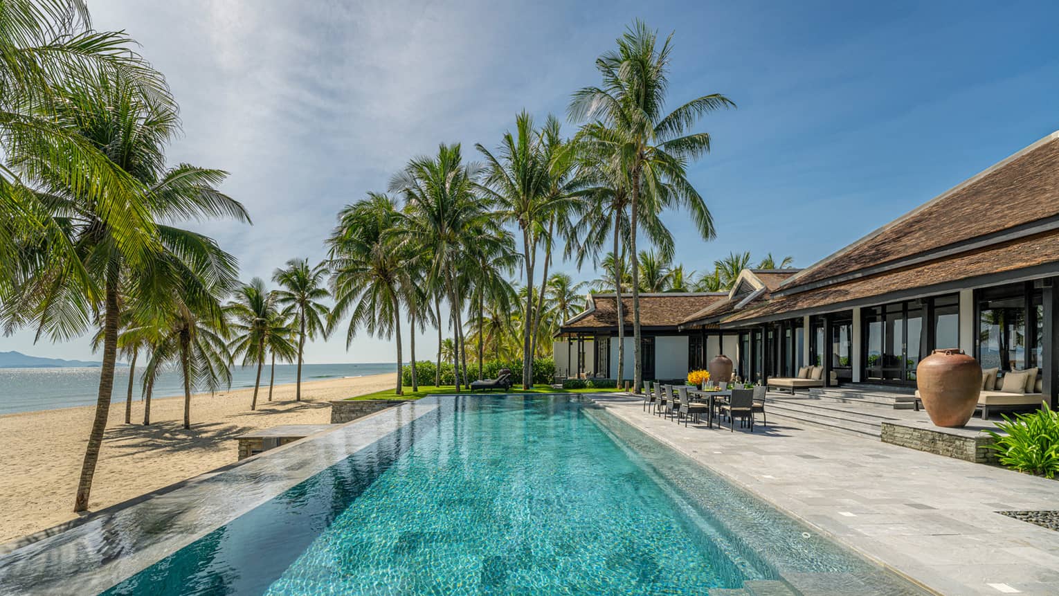 Luxury beachfront villa with a spacious pool surrounded by palm trees, overlooking a sandy beach and calm ocean