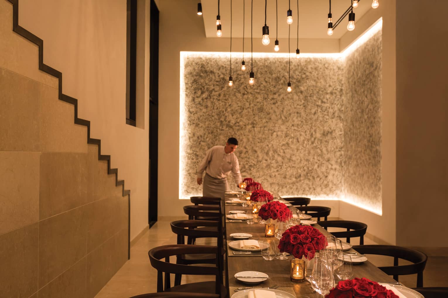 A server lights a candle at the far end of a long table artfully arranged with vases of red roses and set for a large group.