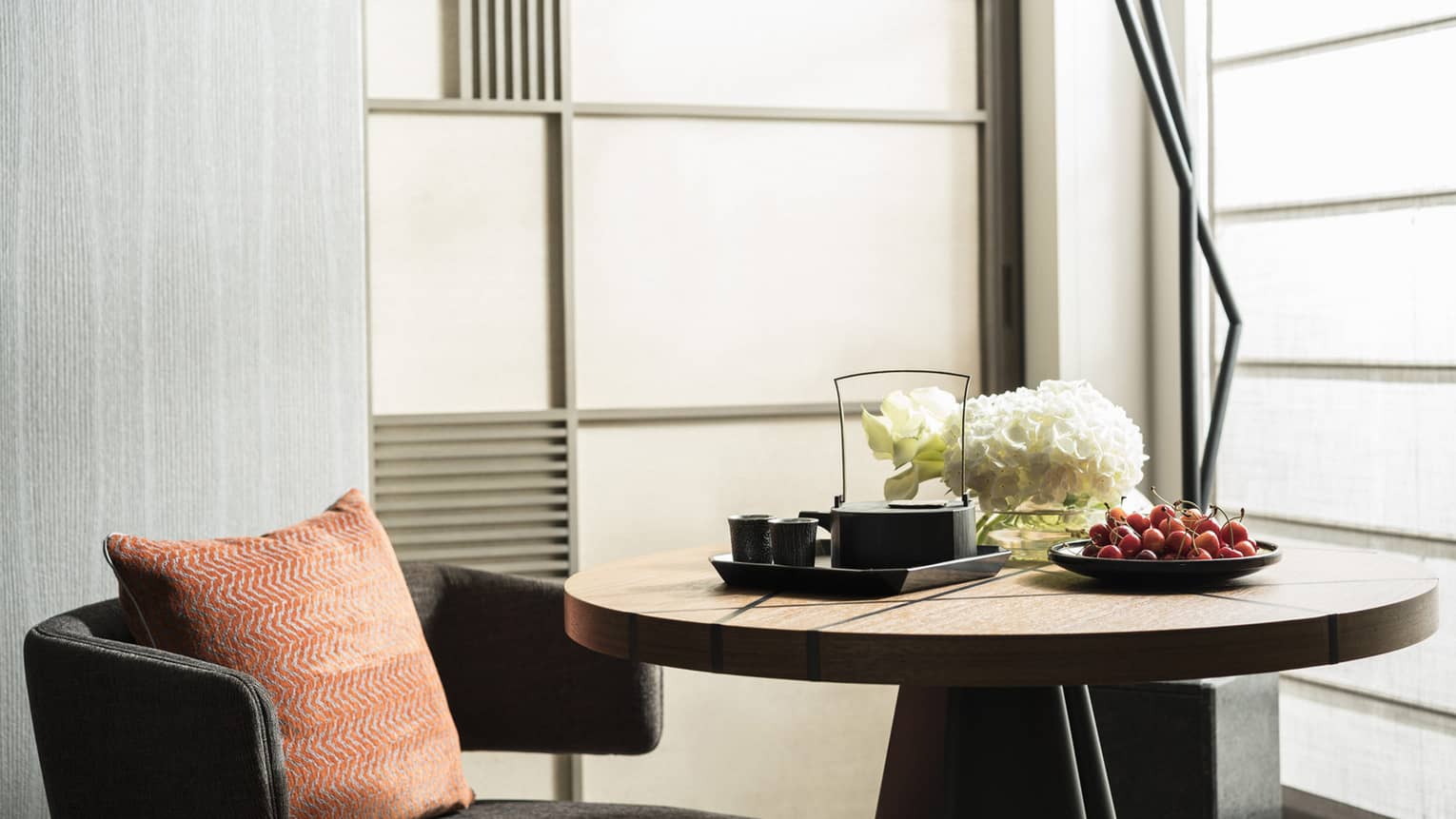 Cozy seating area with a round wooden table set with a tea tray, cherries and white flowers, featuring an orange cushion