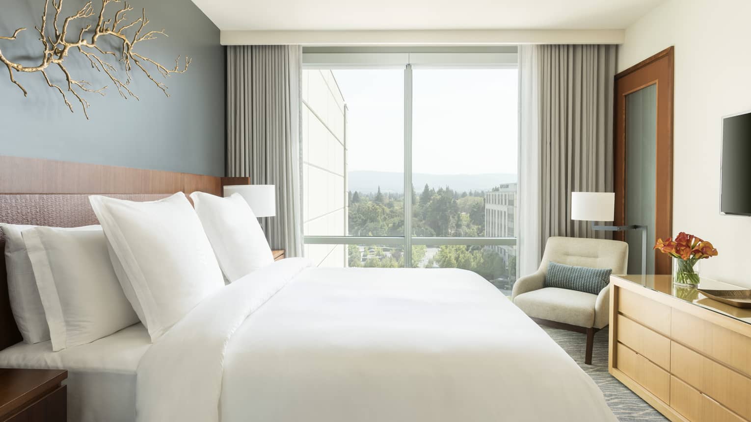 Modern, bright hotel room with gold branch on wall above bed, floor-to-ceiling window