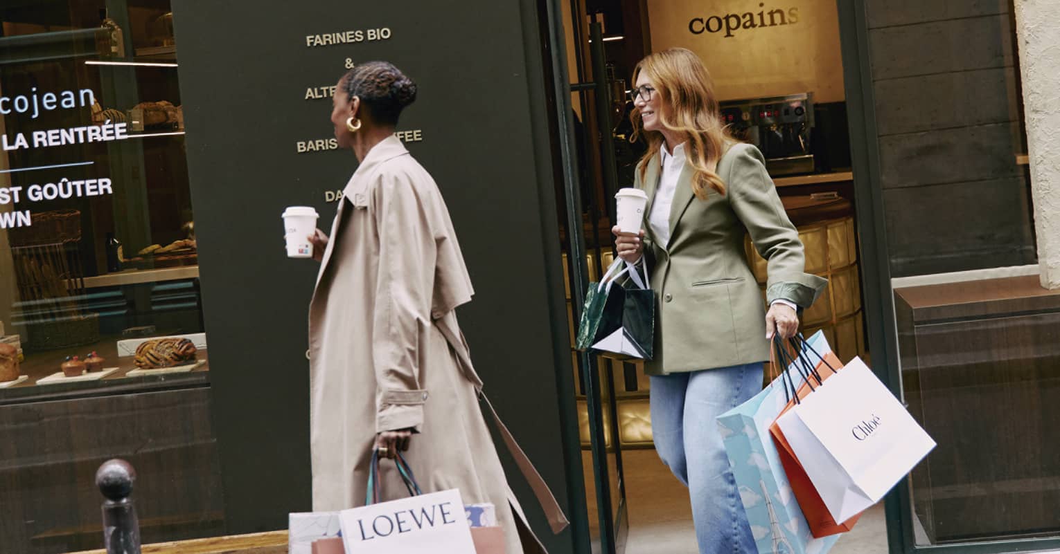 Fashionably dressed tourists with coffee and shopping bags exit an upscale café. Pastries are displayed in the café window.