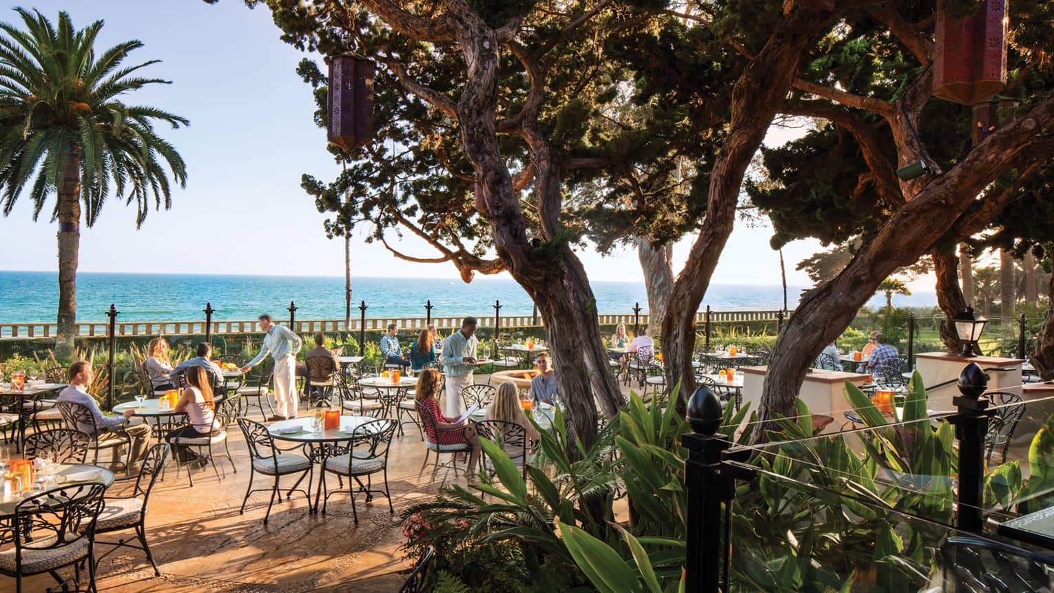 View of diners on Bella Vista patio at wrought-iron tables, chairs through large trees, plants 