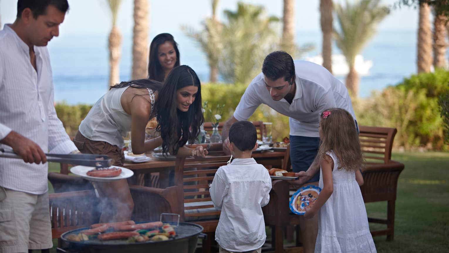 Man and woman dish out dinner for two children by patio table, man flips hot dogs on small barbecue