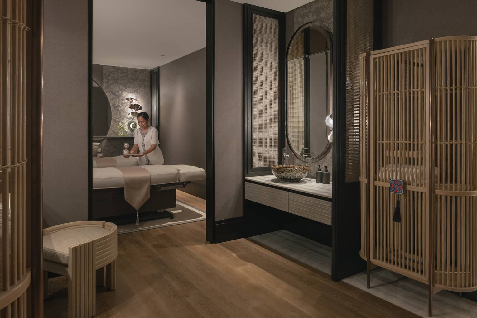 A tray is gently placed on one of two massage tables in a warmly lit spa room with soft grey walls and natural wood décor