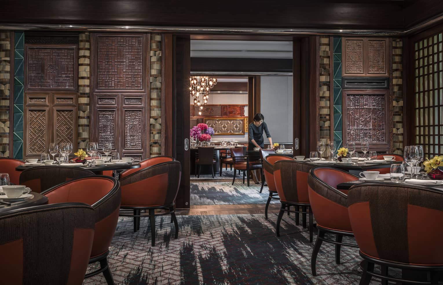 Woman sets table at Jiang-Nan Chun, Michelin-starred Cantonese restaurant, with red leather bucket chairs around dining tables, carved wood walls.