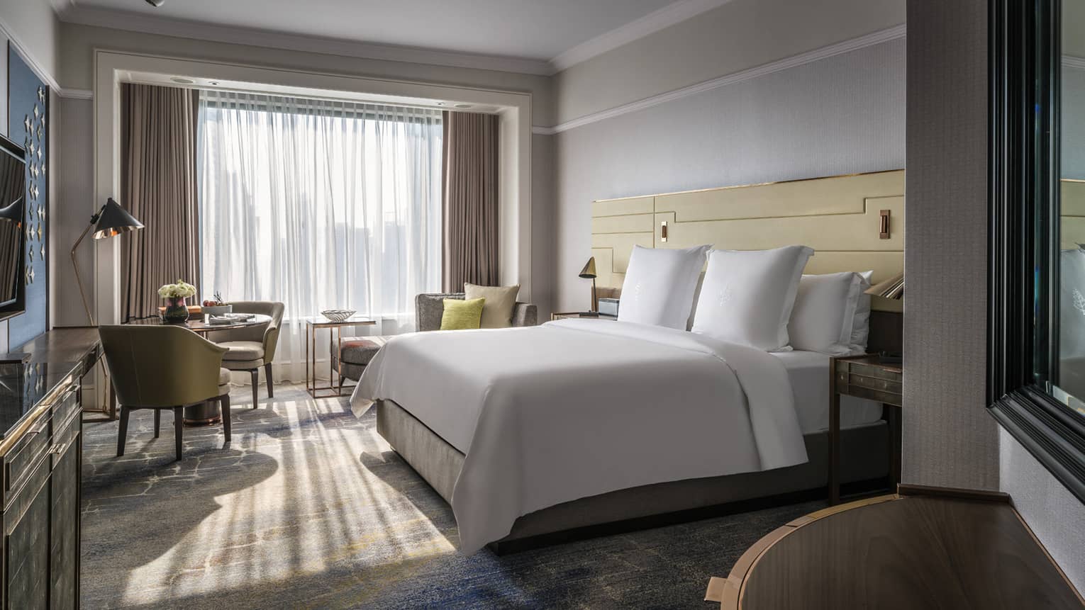 Sunny guest room with bed and white linens, small dining table and chaise lounge, and white curtains