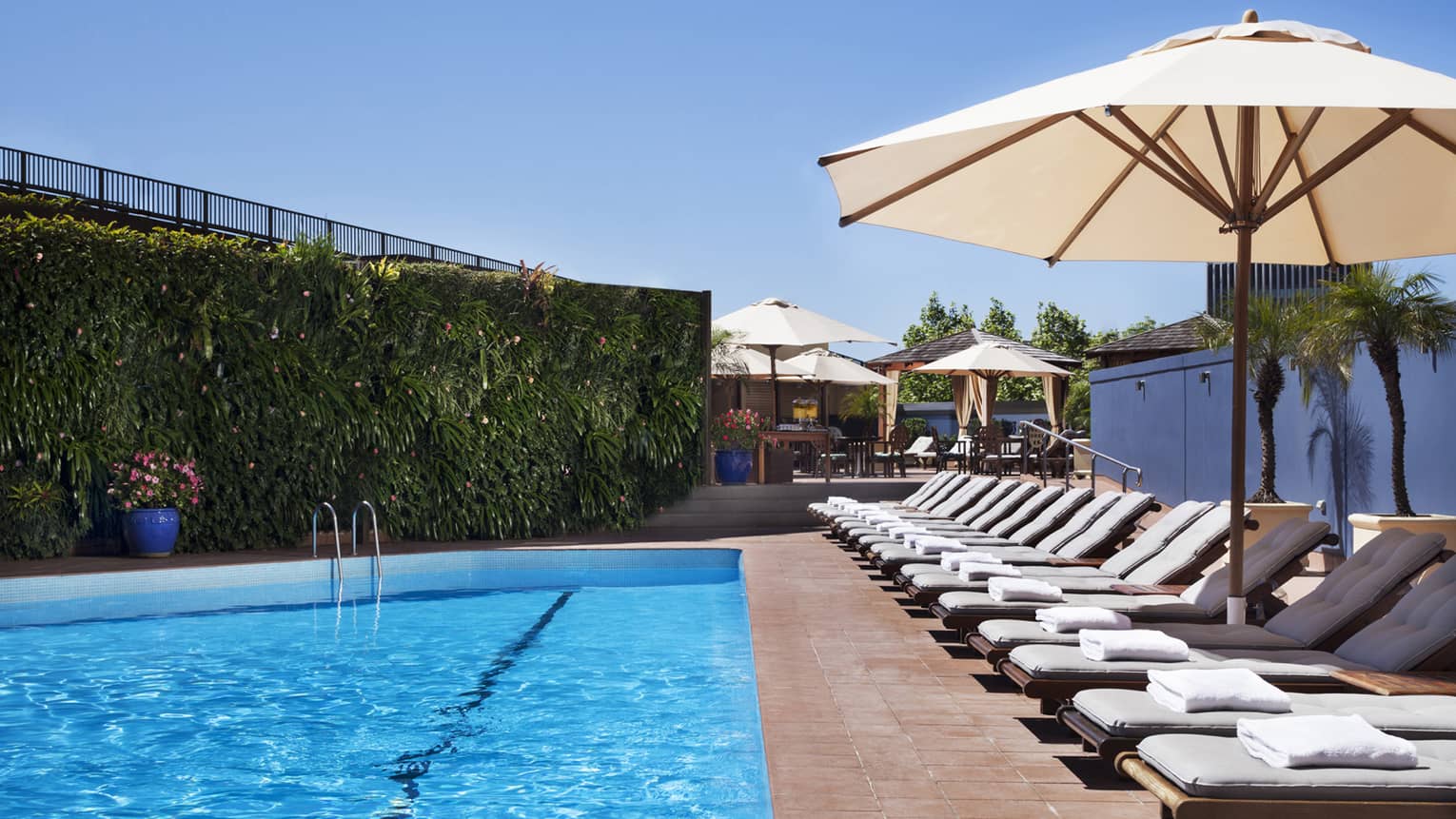 White lounge chairs lined up on deck under patio umbrella by blue outdoor swimming pool