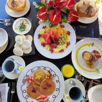 Aerial view of a table filled with white plates containing pancakes, fruits and various breakfast foods, along with two blue coffee mugs and two coconuts with straws