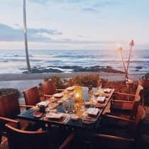 A wooden dining table is set with dishes and lit candles by the beach at sunset