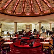 A spacious circular lounge featuring a baby grand piano on a red velvet podium, surrounded by elegant lounge tables